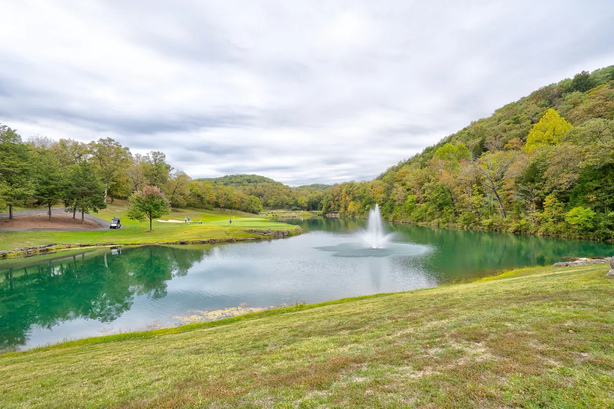 A peaceful view of a calm lake with a fountain, surrounded by rolling hills and greenery. The scene is tranquil, inviting guests to fish, enjoy nature walks, or simply savor the quiet beauty of the outdoors. The changing seasons are reflected in the natural colors and light.