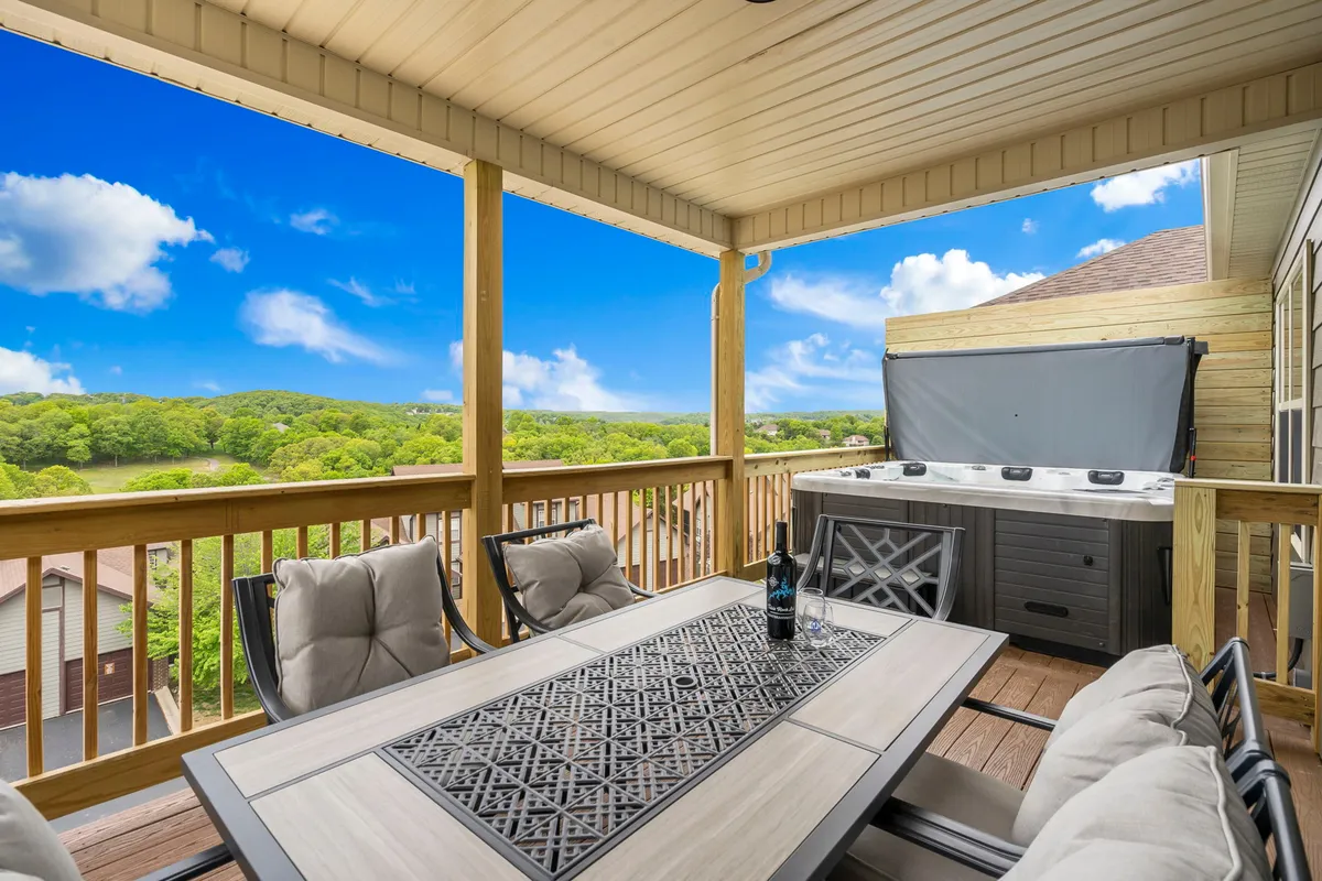 The image showcases the condo's inviting outdoor deck, featuring a stylish dining table set for al fresco meals, a bubbling hot tub, a scenic backdrop of rolling hills, and a bottle of wine ready for guests. The relaxed setting hints at memorable evenings spent watching sunsets, sharing laughter, and enjoying the serenity of the Ozarks.