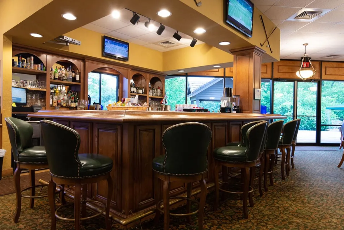 An inviting interior shot of a wooden bar with stools, stocked shelves of bottled drinks, and large windows that suggest a bright, friendly atmosphere. Televisions and relaxed seating imply a perfect spot for post-game refreshments and socializing with friends.