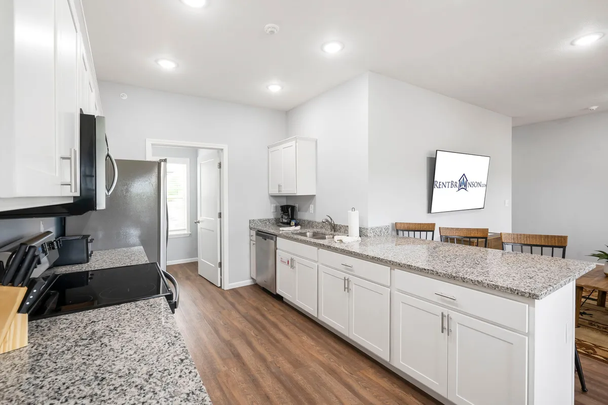 The kitchen is the centerpiece in this image, with a stylish island, granite countertops, stainless steel appliances, and modern cabinetry. A TV screen is visible nearby, suggesting an open relationship between the kitchen and adjacent living areas. The mood is contemporary and inviting, suited for both cooking and entertaining guests.