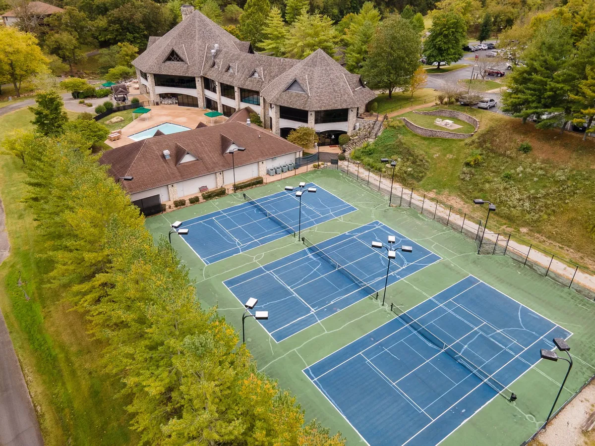 A wide exterior view of tennis courts adjacent to a clubhouse, surrounded by greenery. The courts are ready for action, and the clubhouse stands as a hub for social and group activities.