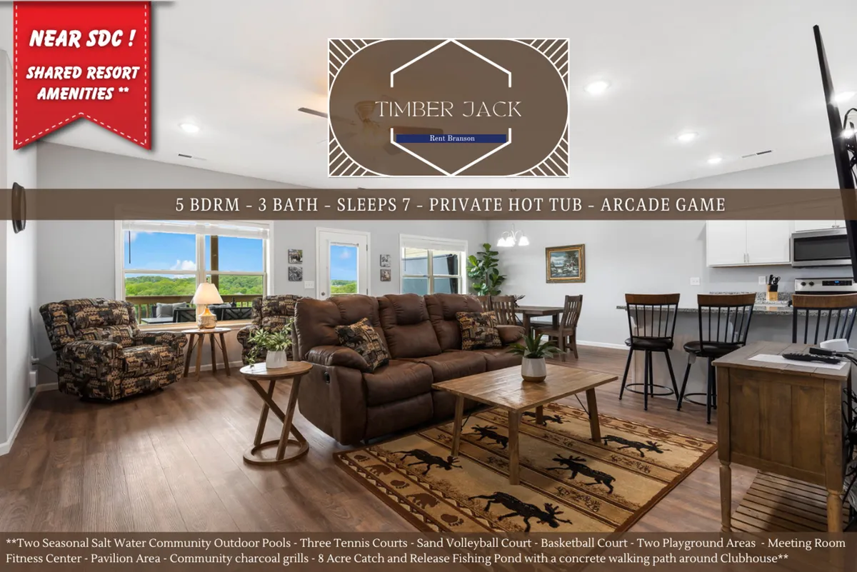 A broad interior view showing a comfortable seating area, large windows with scenic views, a wooden dining table, and part of the kitchen. The space is bright and family-friendly, inviting guests to relax, dine, play games, and soak in the landscape outside.