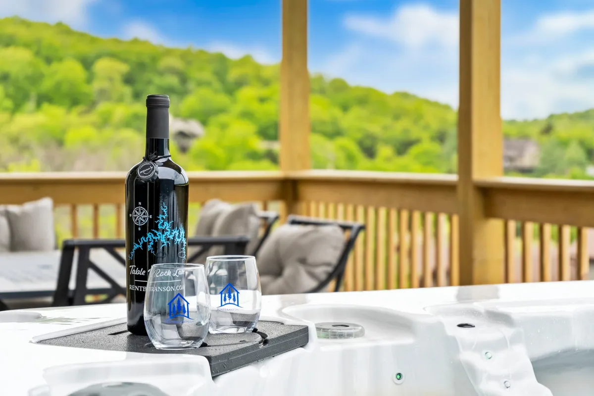 A private outdoor balcony with a bottle of wine and two glasses set beside a hot tub, bordered by a wooden railing and surrounded by lush green hills. The lighting suggests a tranquil, inviting atmosphere ideal for sunset relaxation or a peaceful morning retreat.