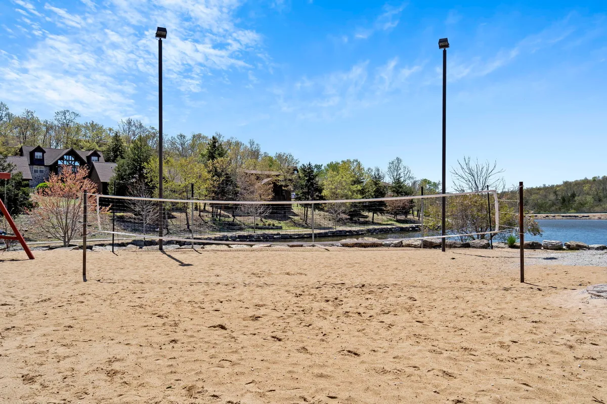 An exterior shot capturing a sand volleyball court set against a scenic lake view, with surrounding greenery and residential buildings visible in the background. The scene is lively yet relaxing, inviting group activities and shared outdoor experiences.