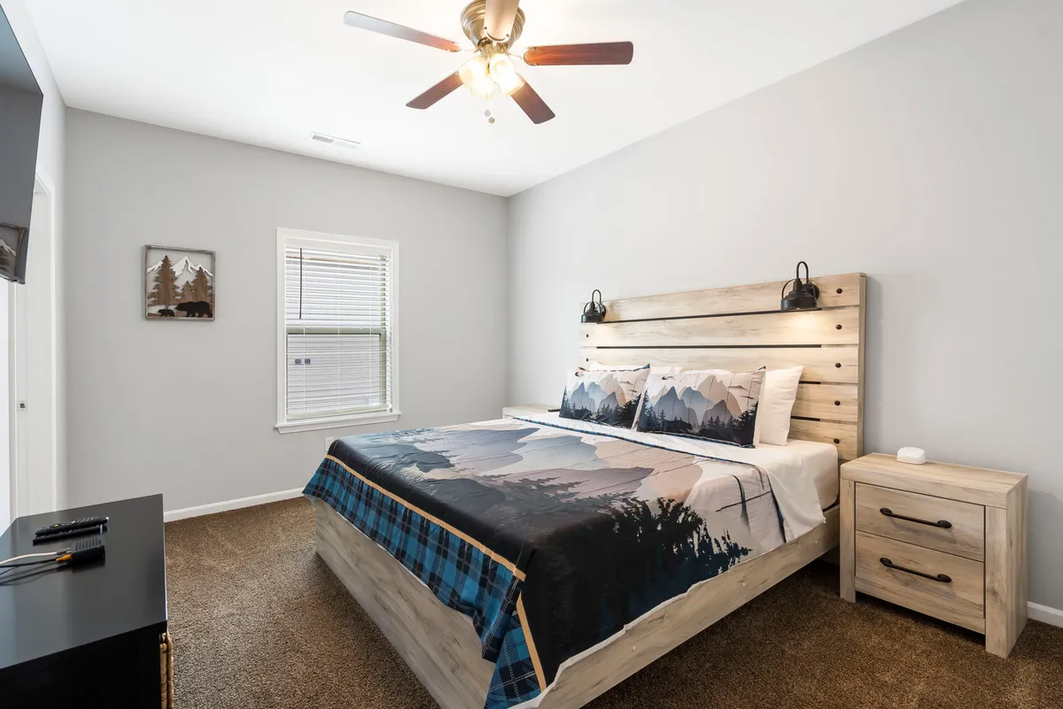 A spacious king bedroom is bathed in sunlight, featuring a large bed dressed in mountain-themed bedding, a cozy nightstand, and tasteful framed artwork. The inviting atmosphere is perfect for relaxation, with an abundance of natural light streaming through the window.
