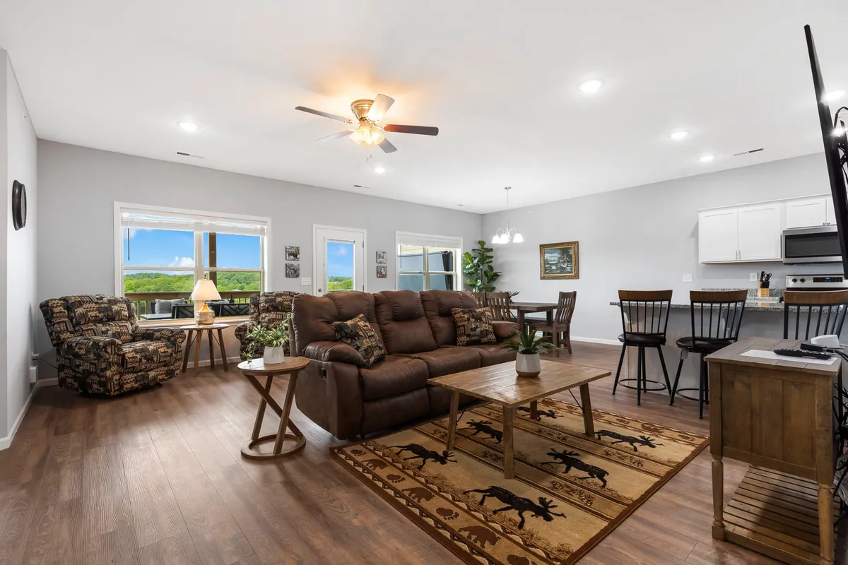 This image depicts a welcoming living area centered around a comfortable brown sofa and a wooden coffee table. The dining area with bar stools is visible in the background, and large windows flood the space with natural light while offering inviting views of the Ozarks. The overall ambiance is cozy and perfect for both relaxation and gatherings.
