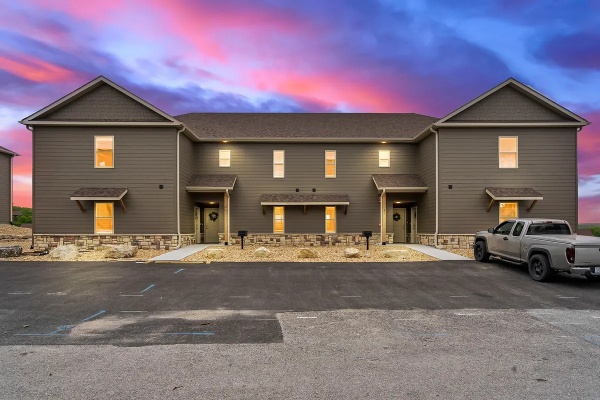 An evening exterior view of a two-story building with warmly lit windows. A vehicle is parked in the foreground, and the setting emanates a welcoming, upscale neighborhood vibe. The porch and communal areas hint at opportunities for socializing and relaxation at the resort.